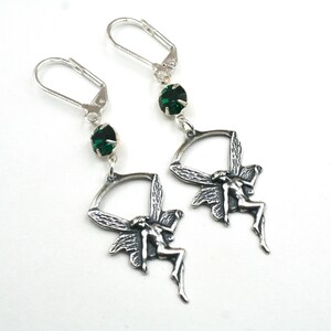 May include: A pair of silver-toned dangle earrings. Each earring features a green gemstone at the top, a curved design, and a detailed fairy figure with outstretched wings. The earrings are set against a white background.