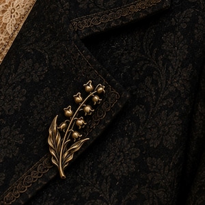 May include: A metallic bronze-toned brooch shaped like a lily of the valley flower with multiple small bell-shaped blossoms and two leaves, pinned to a dark black fabric garment featuring a subtle, intricate tonal floral damask pattern.