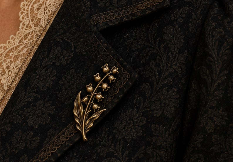May include: A metallic bronze-toned brooch shaped like a lily of the valley flower with multiple small bell-shaped blossoms and two leaves, pinned to a dark black fabric garment featuring a subtle, intricate tonal floral damask pattern.