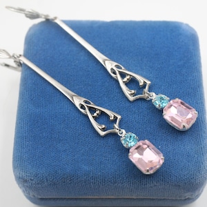 May include: Silver-toned drop earrings with an ornate design. Each earring features a light blue and a pink rectangular gemstone. The earrings are displayed on a blue velvet surface.