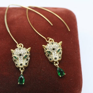 May include: A pair of gold-tone dangle earrings featuring a leopard head design with clear and black rhinestones and a green teardrop-shaped stone hanging from each head.