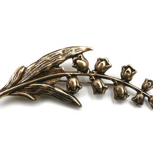 May include: A bronze-toned metal brooch shaped like a lily of the valley plant. The design features a textured, elongated leaf extending to the left and a curved stem on the right adorned with seven small, bell-shaped flower blossoms in a row.
