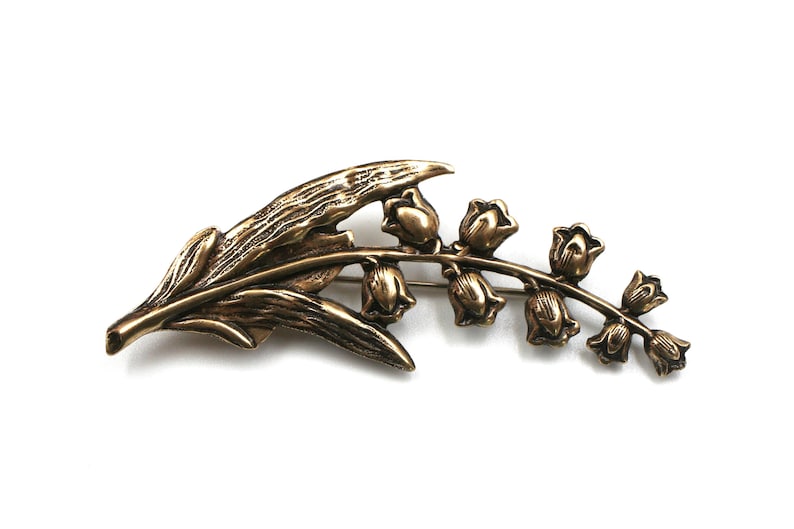 May include: A bronze-toned metal brooch shaped like a lily of the valley plant. The design features a textured, elongated leaf extending to the left and a curved stem on the right adorned with seven small, bell-shaped flower blossoms in a row.