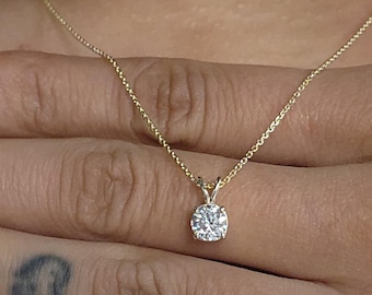 May include: A delicate gold necklace featuring a single, round, clear gemstone pendant. The necklace is displayed on a person's hand, with the focus on the pendant's sparkle. The hand has teal-colored nail polish and a small tattoo.