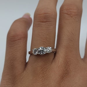May include: A three-stone ring with a gold band and three round, clear gemstones. The ring is displayed on a hand against a white background. The gemstones are of similar size and are set close together.