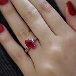 May include: A silver ring with a large, pear-shaped, red gemstone. The hand wearing the ring has red nail polish. A gold chain is visible in the background. The person is wearing a dark gray shirt.