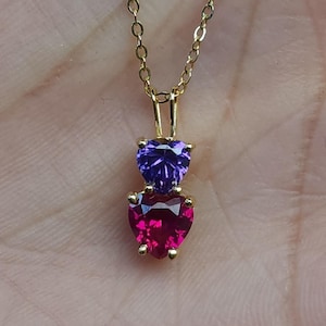 May include: A gold-coloured necklace with a pendant featuring two heart-shaped gemstones. The top gemstone is purple, and the bottom one is a vibrant red. The necklace is displayed against a neutral background.