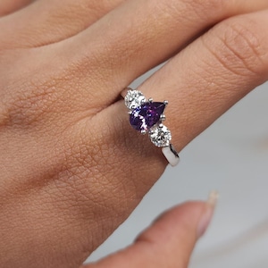 May include: A silver ring featuring a pear-shaped, deep purple gemstone flanked by two smaller, round, clear stones. The ring is set on a hand with a light skin tone, showcasing the jewelry's design and sparkle.