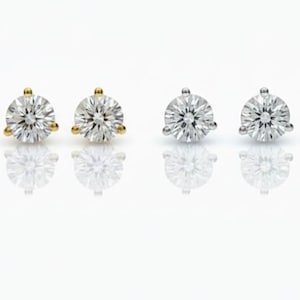 May include: Six pairs of diamond stud earrings in gold, silver, and rose gold settings. Each earring features a round, brilliant-cut diamond. The earrings are displayed on a white surface, with their reflections visible.