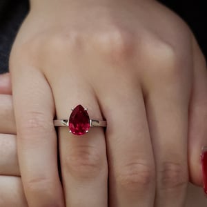 Puede incluir: Un anillo de plata con una piedra roja en forma de pera. El anillo está en el dedo de una persona. Las uñas de la persona están pintadas de rojo.