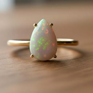 May include: A gold ring featuring a pear-shaped opal gemstone. The opal displays a mix of colors, including green and orange, set against a white background. The ring is set on a wooden surface.