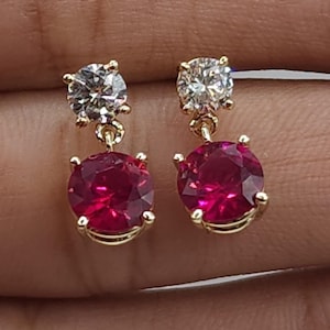May include: A pair of gold earrings with a red and white gemstone design. The earrings feature a round red gemstone with a smaller round white gemstone above it.
