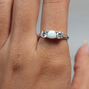 Op de afbeelding: Een zilveren ring met drie stenen. De middelste steen is ovaal en wit, geflankeerd door twee kleinere, ronde, heldere stenen. De ring zit om een vinger en toont het ontwerp. Het is een sieraad.