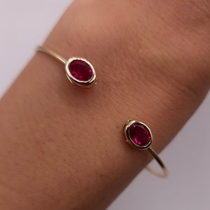 May include: A gold cuff bracelet with two oval, deep red gemstones. The bracelet has a simple, open design, with the gemstones set in a bezel setting. The bracelet is shown on a wrist.