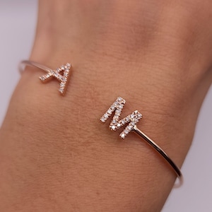 Puede incluir: Brazalete abierto de oro rosa con las letras "A" y "M", cada una incrustada con pequeños cristales transparentes brillantes. El brazalete se muestra en una muñeca, destacando el diseño delicado y el estilo personalizado.