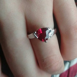 K&ouml;nnte beinhalten: Ein silberner Ring mit einem herzf&ouml;rmigen, tiefroten Edelstein. Der Ring hat ein schlichtes Band und kleine Akzentsteine auf beiden Seiten des Herzens. Der Edelstein ist facettiert, f&auml;ngt das Licht ein und erzeugt einen funkelnden Effekt.