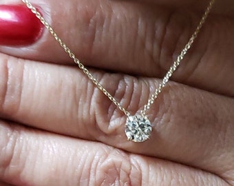 May include: A delicate gold necklace featuring a single, round, clear gemstone pendant. The necklace is displayed against a background of a patterned black and white top and a hand with red nail polish.