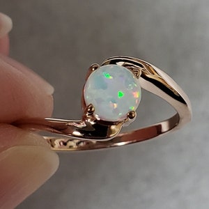 May include: A rose gold ring featuring a round, white opal gemstone. The opal has flashes of green and other colors. The ring's band curves around the stone in a modern, elegant design.