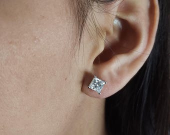May include: A close-up of a person's ear with a square-cut diamond stud earring. The earring is set in a silver or white metal setting. The diamond appears to be clear and sparkling.
