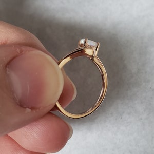 May include: A rose gold ring with a rectangular, white gemstone. The band curves gracefully, leading to the gemstone setting. The ring is held against a soft, neutral background, highlighting its elegant design.