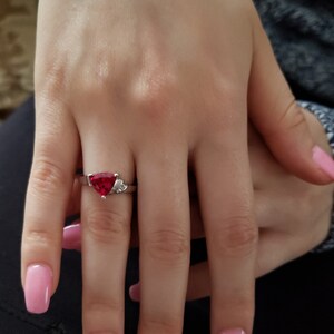 K&ouml;nnte beinhalten: Ein silberner Ring mit einem gro&szlig;en, dreieckigen roten Edelstein und kleinen Akzentsteinen. Der Ring befindet sich an einem Finger mit rosa Nagellack. Die Hand ruht auf einer dunklen Oberfl&auml;che.