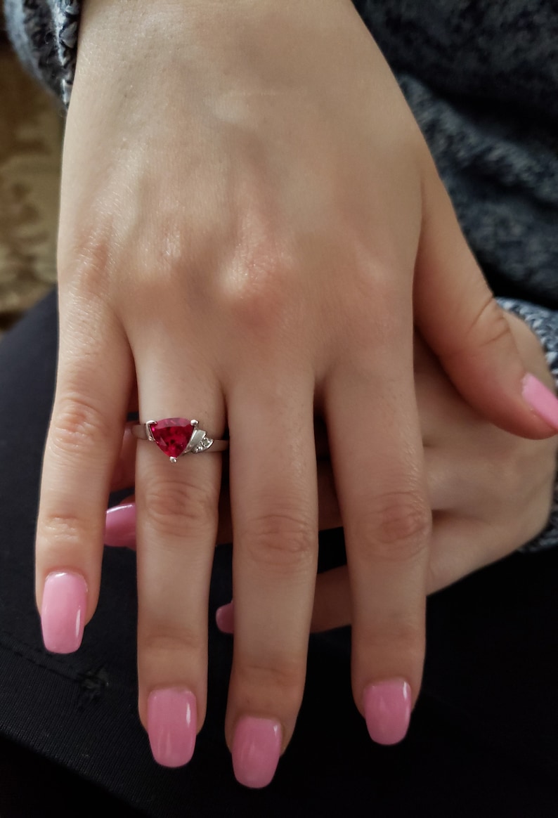 K&ouml;nnte beinhalten: Ein silberner Ring mit einem gro&szlig;en, dreieckigen roten Edelstein und kleinen Akzentsteinen. Der Ring befindet sich an einem Finger mit rosa Nagellack. Die Hand ruht auf einer dunklen Oberfl&auml;che.