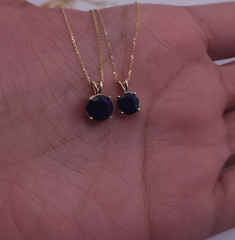 May include: Two gold necklaces with blue gemstone pendants. The necklaces are on a hand.
