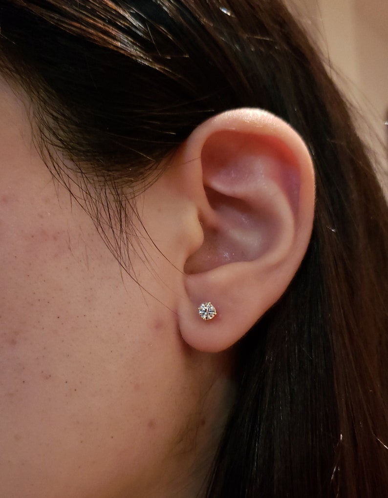May include: A close-up of a person's ear with a small, round, clear diamond stud earring.