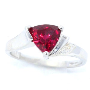 K&ouml;nnte beinhalten: Ein silberner Ring mit einem gro&szlig;en, dreieckigen, tiefroten Edelstein. Das Band ist glatt und poliert, mit kleinen Akzentsteinen an der Seite des Edelsteins. Der Ring ist vor einem wei&szlig;en Hintergrund platziert.