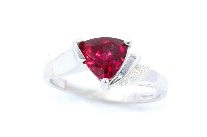 K&ouml;nnte beinhalten: Ein silberner Ring mit einem gro&szlig;en, dreieckigen, tiefroten Edelstein. Das Band ist glatt und poliert, mit kleinen Akzentsteinen an der Seite des Edelsteins. Der Ring ist vor einem wei&szlig;en Hintergrund platziert.