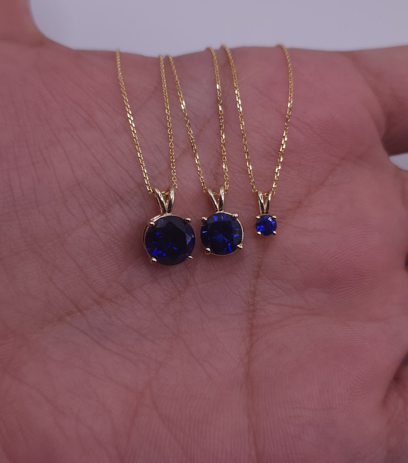 May include: Three gold necklaces with blue gemstone pendants of different sizes. The necklaces are on a hand, with the pendants facing the camera.