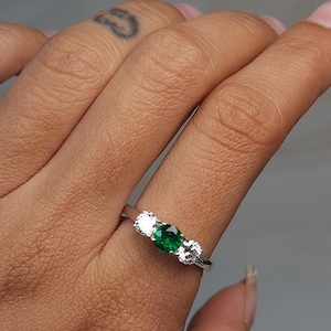 Pode incluir: Um anel de prata com uma pedra central de esmeralda oval e duas pedras redondas e transparentes. O anel está num dedo, destacando o seu design e brilho. As pedras são engastadas numa banda de prata.