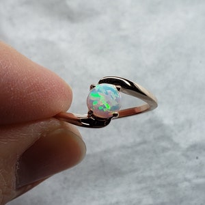May include: Rose gold ring featuring a round opal gemstone with iridescent green and blue flashes. The ring has a delicate, swirling design that cradles the opal. The gemstone is approximately 0.3 inches in diameter.