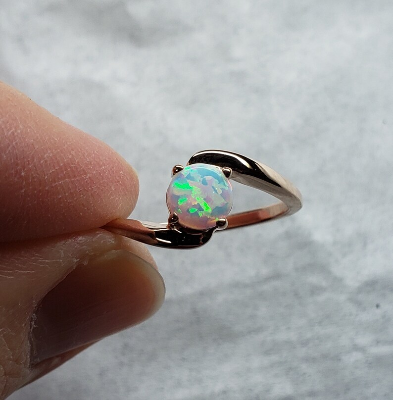May include: Rose gold ring featuring a round opal gemstone with iridescent green and blue flashes. The ring has a delicate, swirling design that cradles the opal. The gemstone is approximately 0.3 inches in diameter.