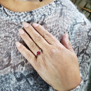 K&ouml;nnte beinhalten: Ein silberner Ring mit einem birnenf&ouml;rmigen, tiefroten Edelstein und kleinen Akzentsteinen. Der Ring befindet sich an einer Hand mit einem hellen Pullover und einer silbernen Halskette mit schwarzen Perlen.
