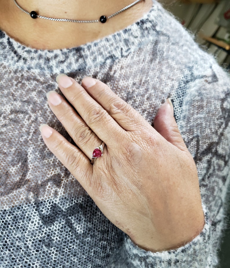 K&ouml;nnte beinhalten: Ein silberner Ring mit einem birnenf&ouml;rmigen, tiefroten Edelstein und kleinen Akzentsteinen. Der Ring befindet sich an einer Hand mit einem hellen Pullover und einer silbernen Halskette mit schwarzen Perlen.