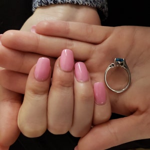 K&ouml;nnte beinhalten: Ein silberner Ring mit einem rechteckigen, blauen Edelstein wird in einer Hand mit rosa Nagellack gehalten. Der Ring hat ein schlichtes Band und kleine Akzentsteine. Die N&auml;gel sind hellrosa lackiert.
