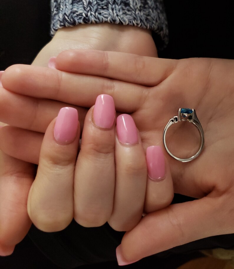 K&ouml;nnte beinhalten: Ein silberner Ring mit einem rechteckigen, blauen Edelstein wird in einer Hand mit rosa Nagellack gehalten. Der Ring hat ein schlichtes Band und kleine Akzentsteine. Die N&auml;gel sind hellrosa lackiert.