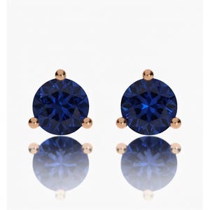 May include: A pair of stud earrings featuring a deep blue, round gemstone set in a rose gold metal. Each earring has three small rose gold accents around the central stone. The earrings are displayed on a reflective surface.