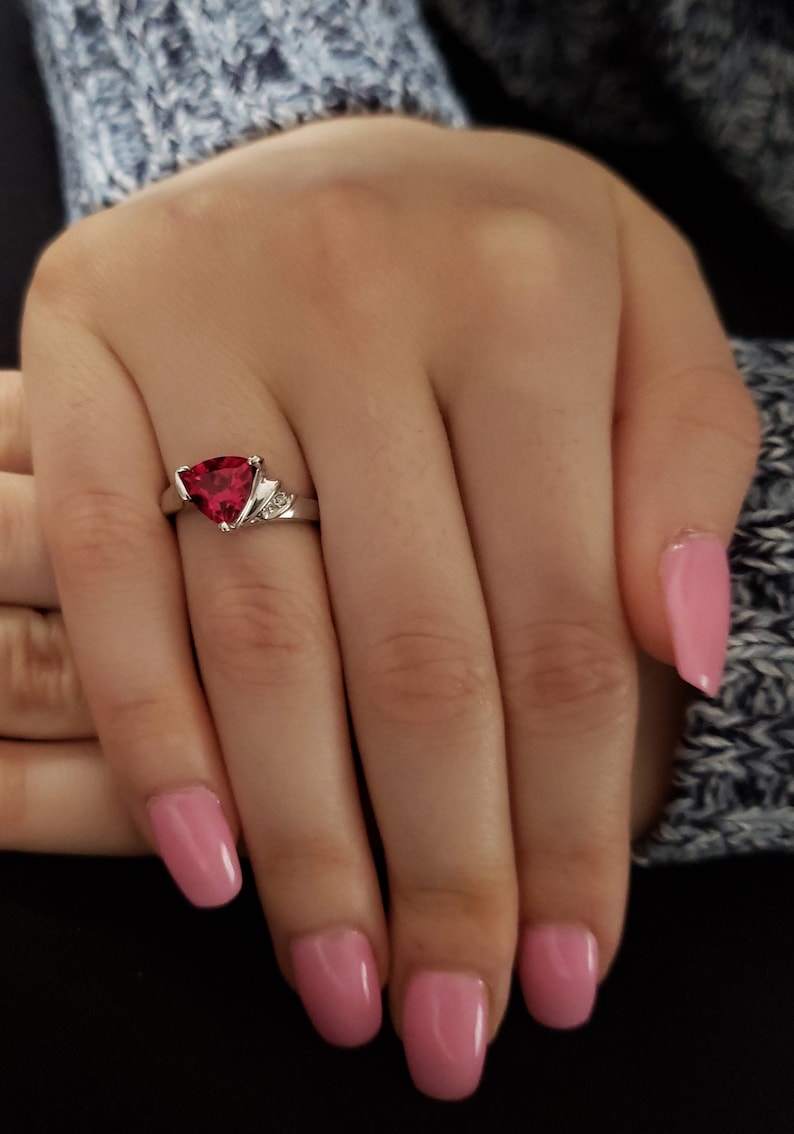 K&ouml;nnte beinhalten: Ein silberner Ring mit einem dreieckigen, tiefroten Edelstein. Der Ring hat kleine Akzentsteine auf beiden Seiten des Hauptsteins. Die Hand, die den Ring tr&auml;gt, hat rosa Nagellack und liegt auf einem blau-grauen Pullover.