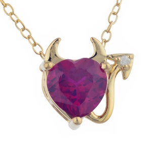 May include: A gold-colored necklace featuring a heart-shaped, deep red gemstone pendant. The pendant is framed by a gold design with small horns and a diamond accent. The chain is also gold.