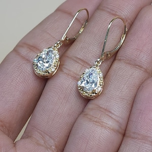 Puede incluir: Un par de pendientes colgantes dorados. Cada pendiente presenta una piedra preciosa clara en forma de pera, rodeada de piedras preciosas claras más pequeñas. Los pendientes se muestran sobre un fondo neutro.