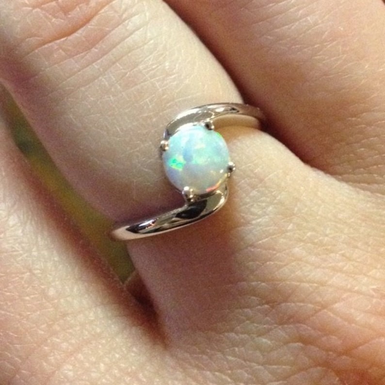May include: A silver ring with a round, iridescent opal gemstone. The opal is held in place by four prongs. The ring's band curves around the opal in a stylish design. The ring is shown on a finger.
