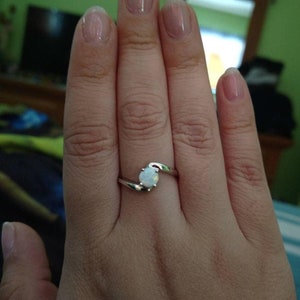 May include: A silver ring with a white opal gemstone. The ring features a swirling design where the band curves to cradle the oval-shaped opal. The ring is shown on a hand.