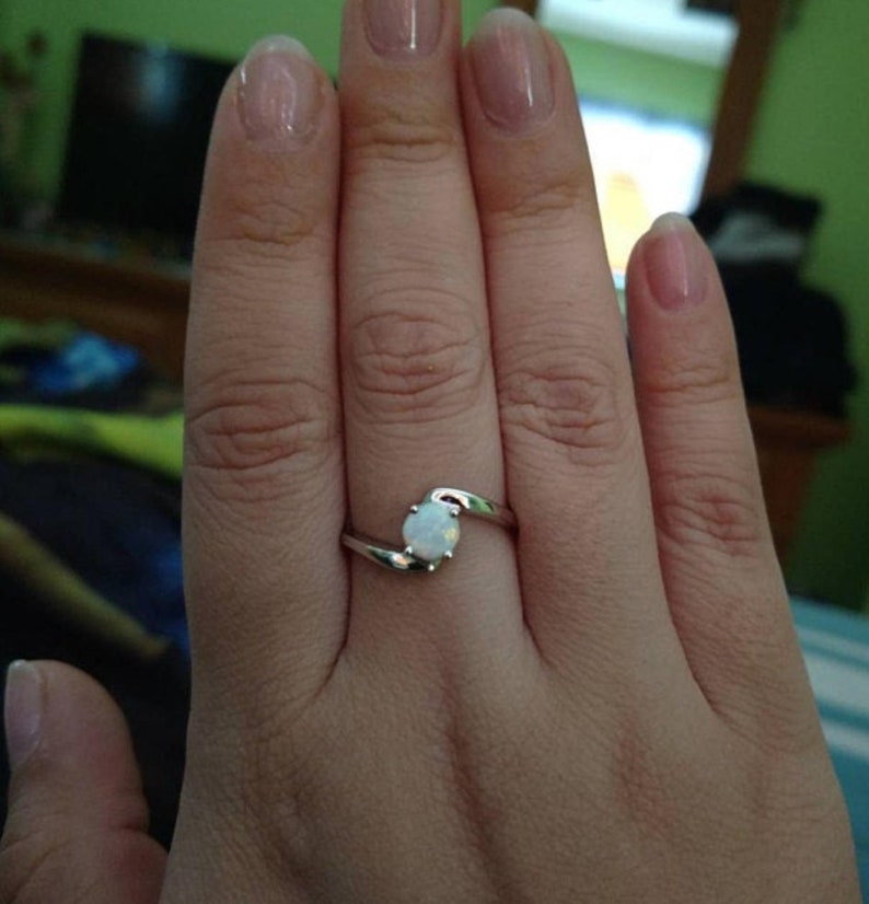May include: A silver ring with a white opal gemstone. The ring features a swirling design where the band curves to cradle the oval-shaped opal. The ring is shown on a hand.