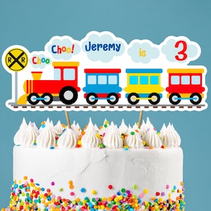 Op de afbeelding: Een witte cake met kleurrijke sprinkels, bedekt met een trein-taarttopper. De topper heeft een rode, gele en blauwe trein met de woorden "Choo! Choo! Jeremy is 3" erop.