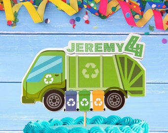 May include: A green and blue garbage truck cake topper with the name "JEREMY" and the number "4" on it. The truck has recycling bins on the back and is on top of a blue and white frosted cake with colorful sprinkles.