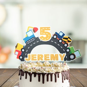 May include: A birthday cake with a construction vehicle themed cake topper. The topper features a yellow number 5, a road, and several construction vehicles. The cake is decorated with chocolate frosting and red and white sprinkles. The cake topper says "JEREMY".
