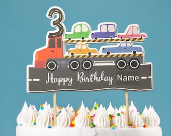 May include: A cake topper featuring a red truck carrying colorful cars, with a banner that reads "Happy Birthday Name".