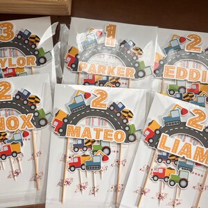 May include: Six construction-themed cake toppers with yellow, orange, and blue accents. Each topper features a number, a name, and a road with construction vehicles. The names are Baylor, Parker, Eddie, Knox, Mateo, and Liam.